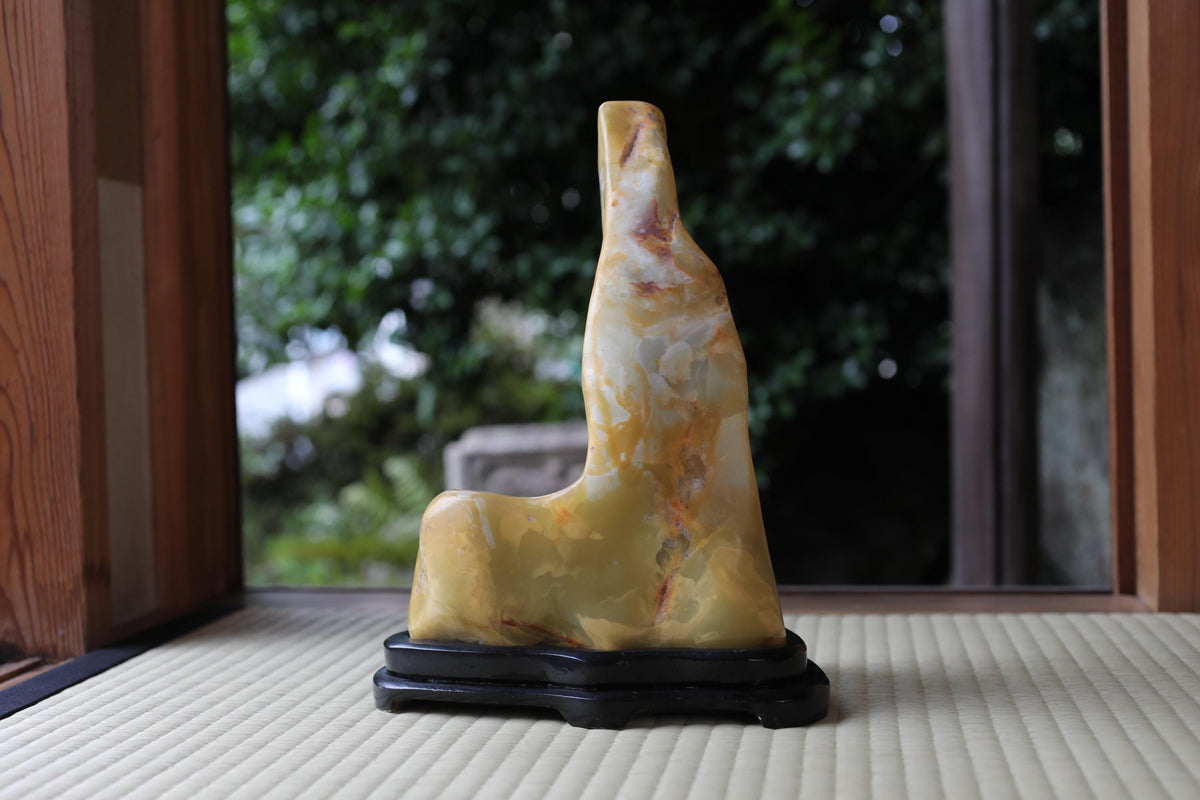 A yellow and orange stone sculpture is displayed on a black stand, positioned in front of a window with a view of trees.