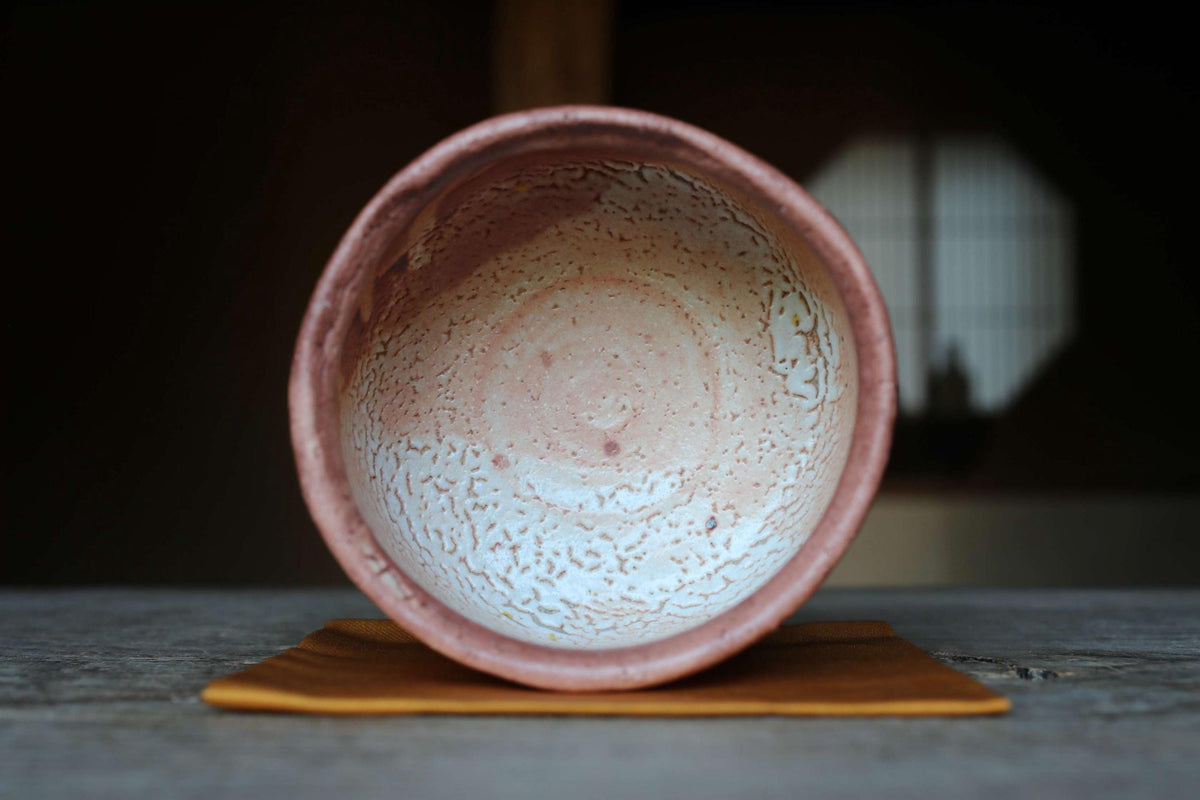 A ceramic bowl with a pinkish-brown hue sits on a brown cloth, with a blurred background that appears to be a wooden structure.
