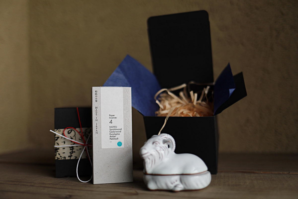 a ceramic ram figurine, a black box with a blue lid, and a white paper bag with text on it, all placed on a wooden surface.