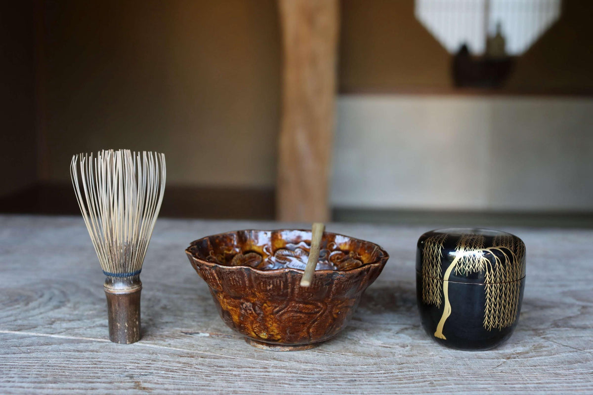 three ceramic objects arranged on a wooden surface: a metal whisk, a bowl, and a small black container with a gold design.