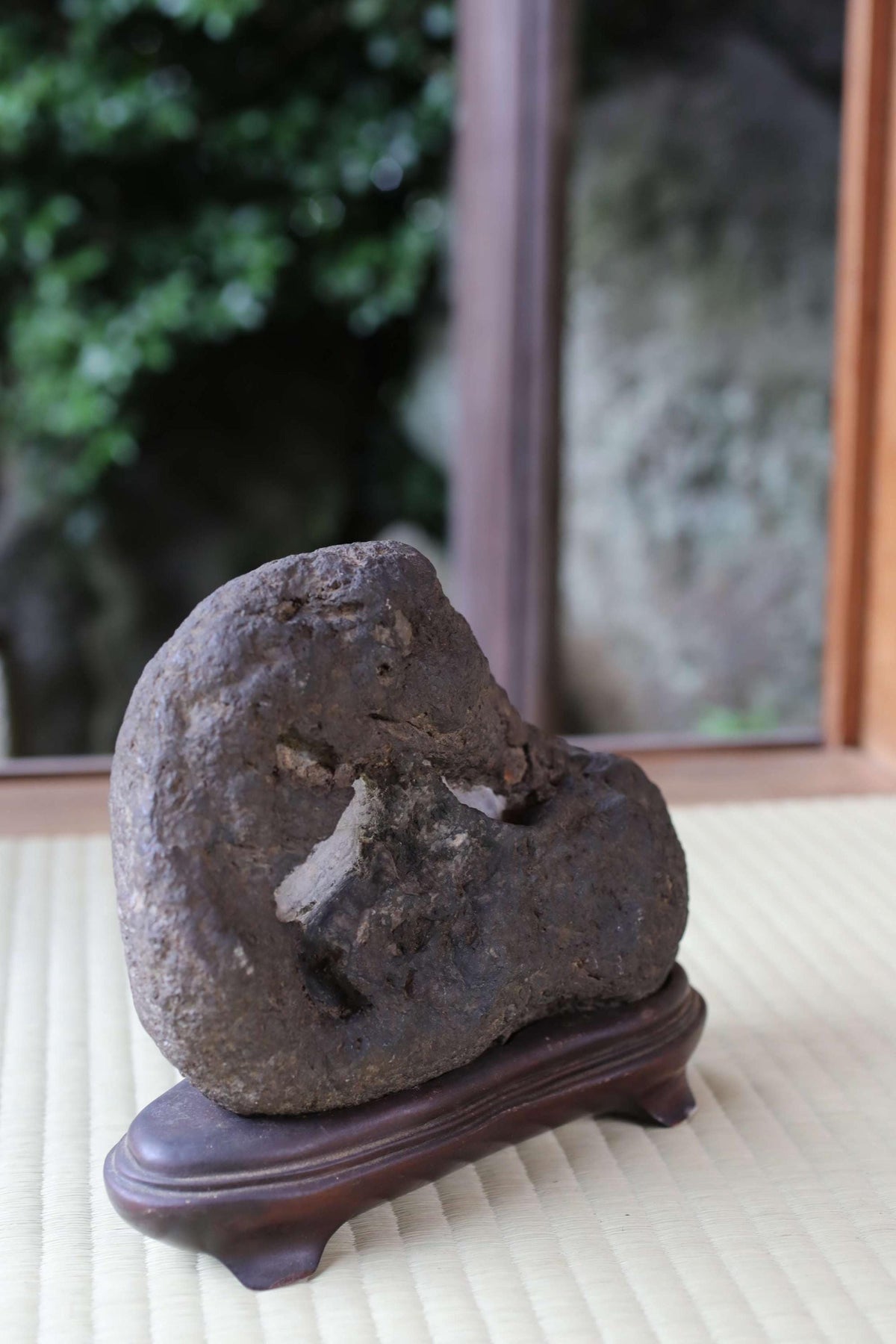 A large, dark rock sits on a wooden stand in front of a window, with a blurred natural background.