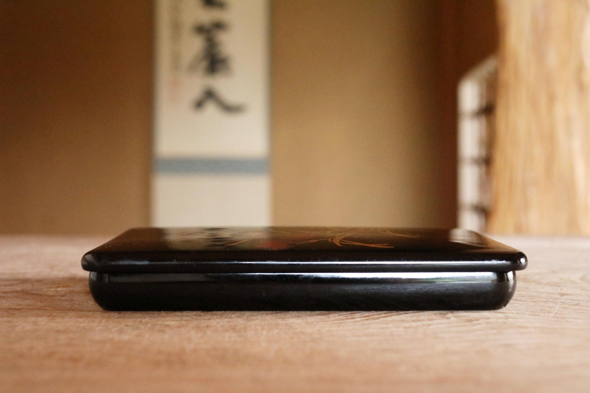 A black lacquer box with a decorative design sits on a wooden surface, with a blurred background that includes a wall with Japanese characters.