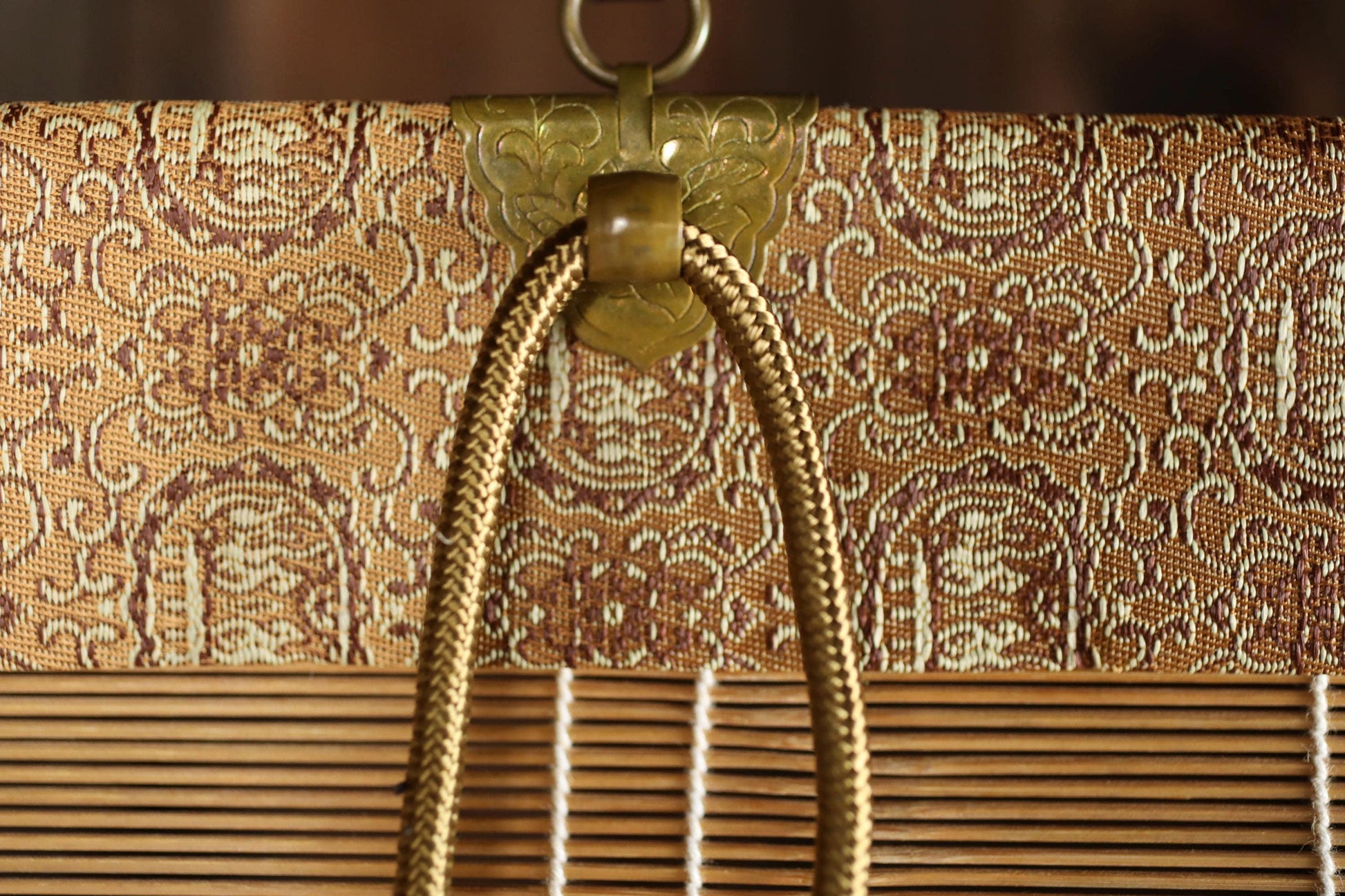 a close-up view of a decorative metal clasp and a woven bamboo blind, with a gold-colored rope hanging from the clasp.