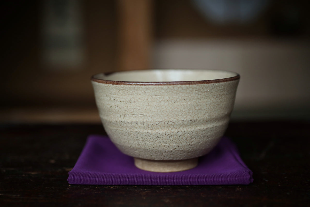 A white ceramic bowl with a brown rim sits on a purple cloth on a wooden surface.