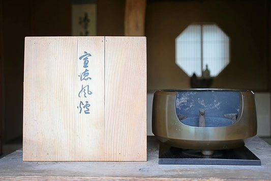 a wooden box with Japanese characters on it, placed next to a metal object, possibly a bowl or container, on a wooden surface.