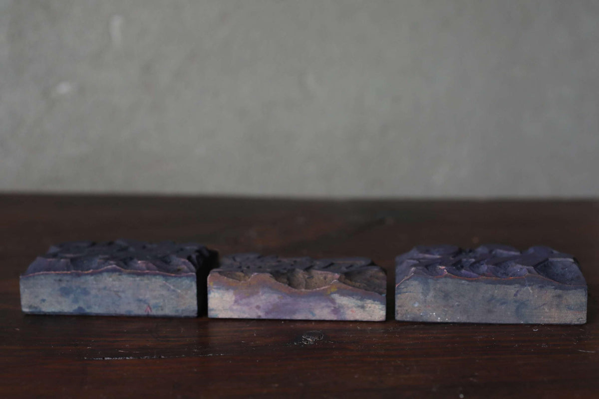 Three rectangular blocks of varying shades of purple are arranged on a wooden surface against a plain background.