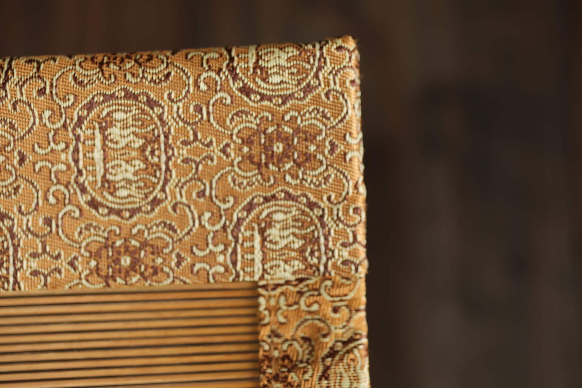 a close-up view of a decorative fabric with a repeating pattern of swirls and lines in shades of brown and beige. The fabric appears to be a piece of upholstery or a textile with an intricate design. The background is blurred, drawing attention to the fabric and its details.