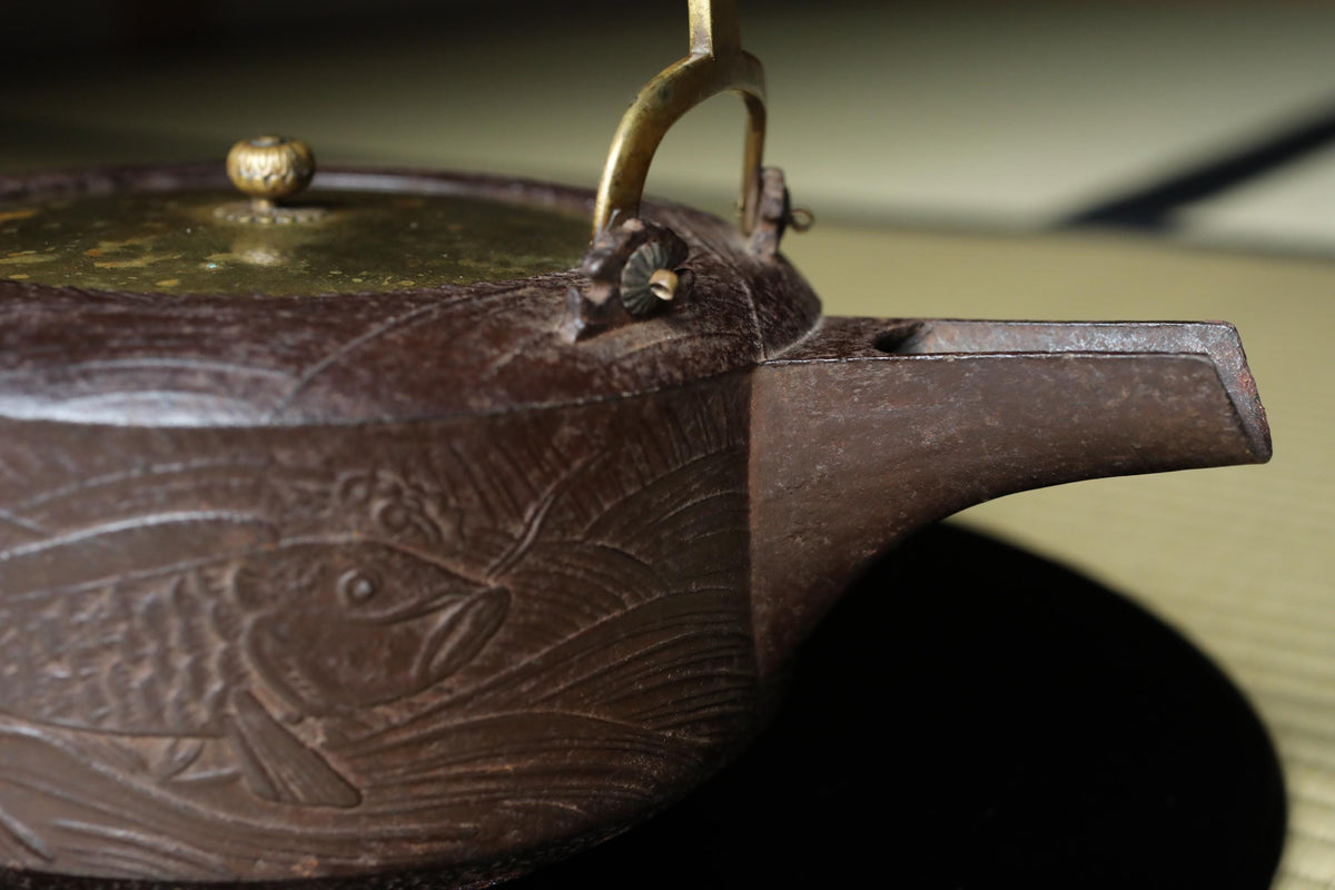 A close-up image of a brown, antique-looking tea kettle with a fish design on it, resting on a surface.