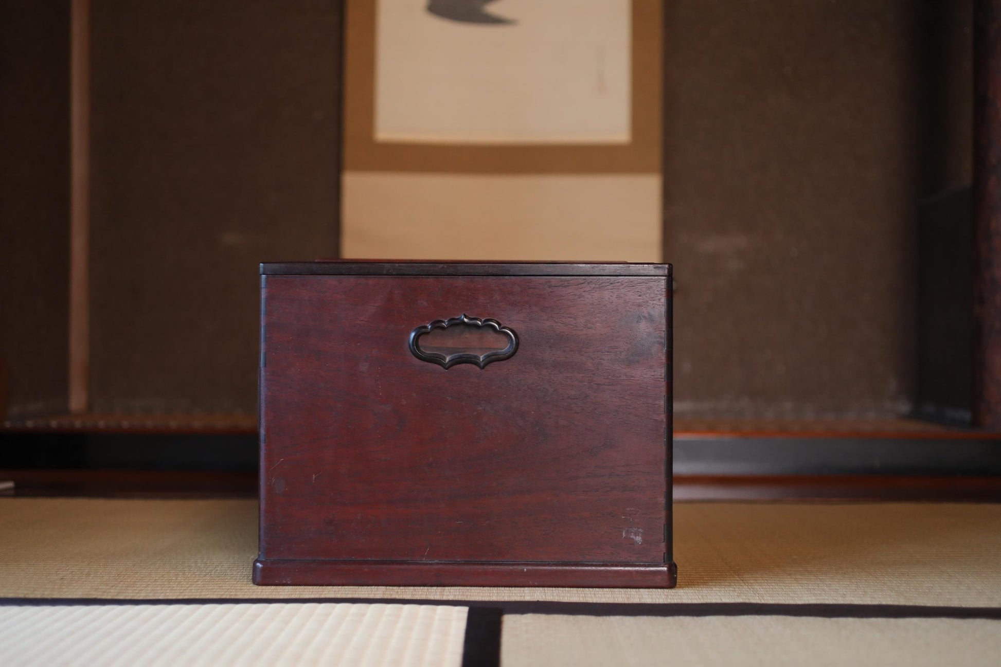 A dark wooden box with a handle on top, placed on a beige mat in front of a wall with a picture frame.