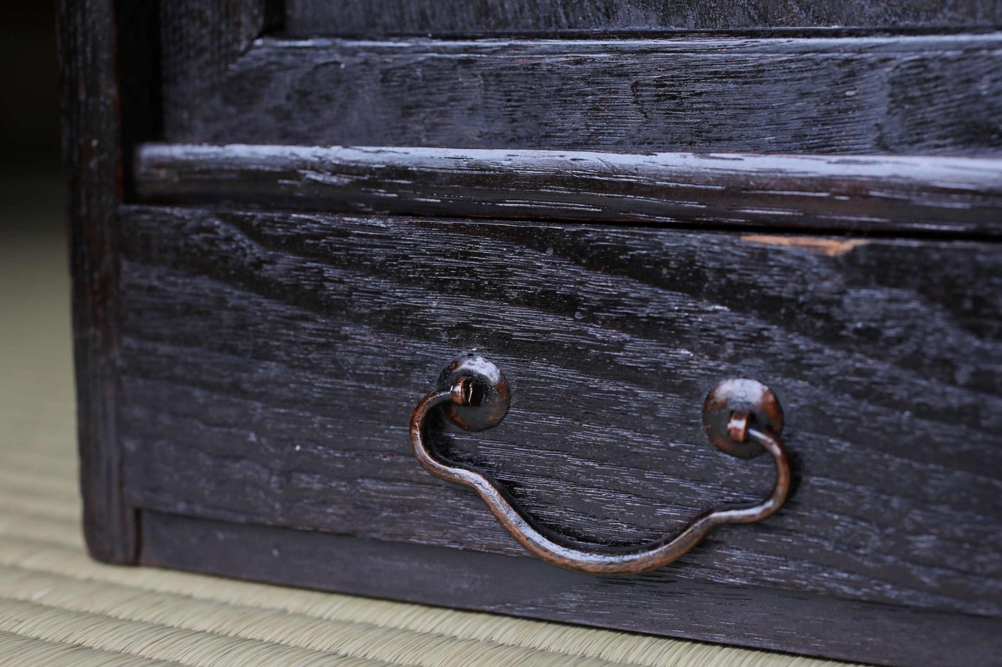 A close-up view of a dark wooden drawer with a curved metal handle, set against a blurred background.