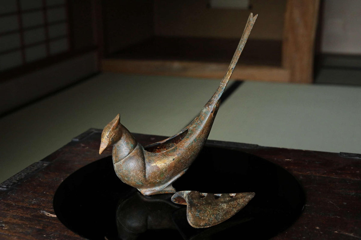 An antique bronze bird figurine is displayed on a wooden surface, with a reflection visible in the surface below.