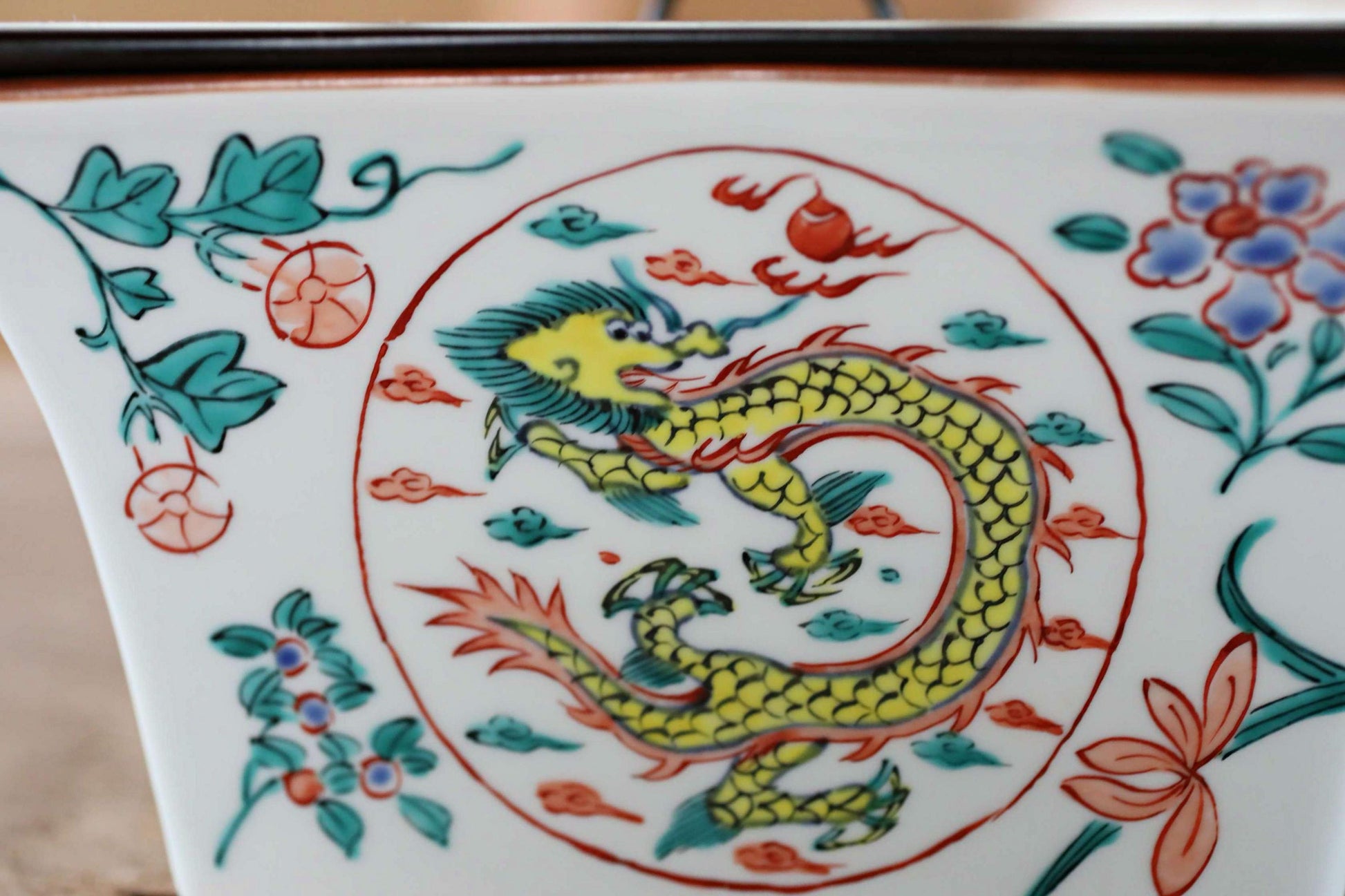 a close-up view of a decorative ceramic piece with a vibrant dragon design. The dragon is depicted in a dynamic pose, surrounded by various colorful elements such as flowers, leaves, and other decorative motifs. The overall composition of the image is visually striking and captures the intricate details of the ceramic artwork.