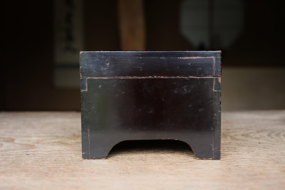 A black wooden box with a weathered appearance sits on a wooden surface.
