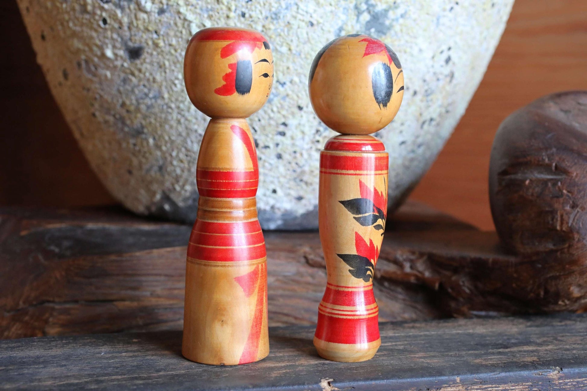 Two wooden dolls with red and black designs stand on a wooden surface, with a large stone vase in the background.