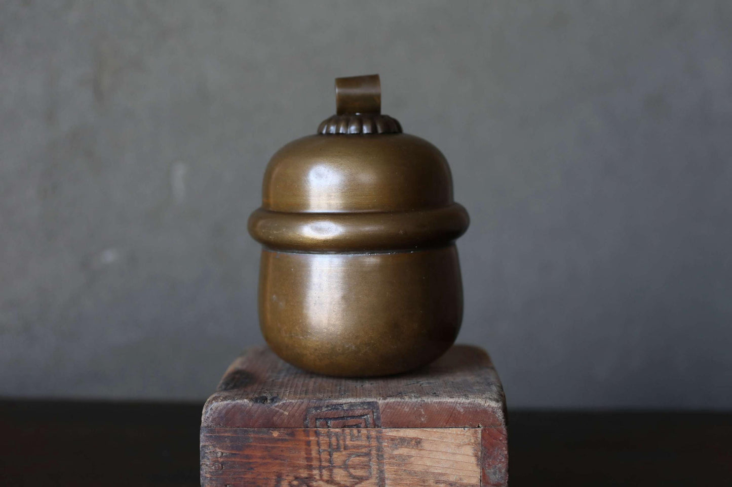 A brass container with a lid sits on a wooden block against a gray background.