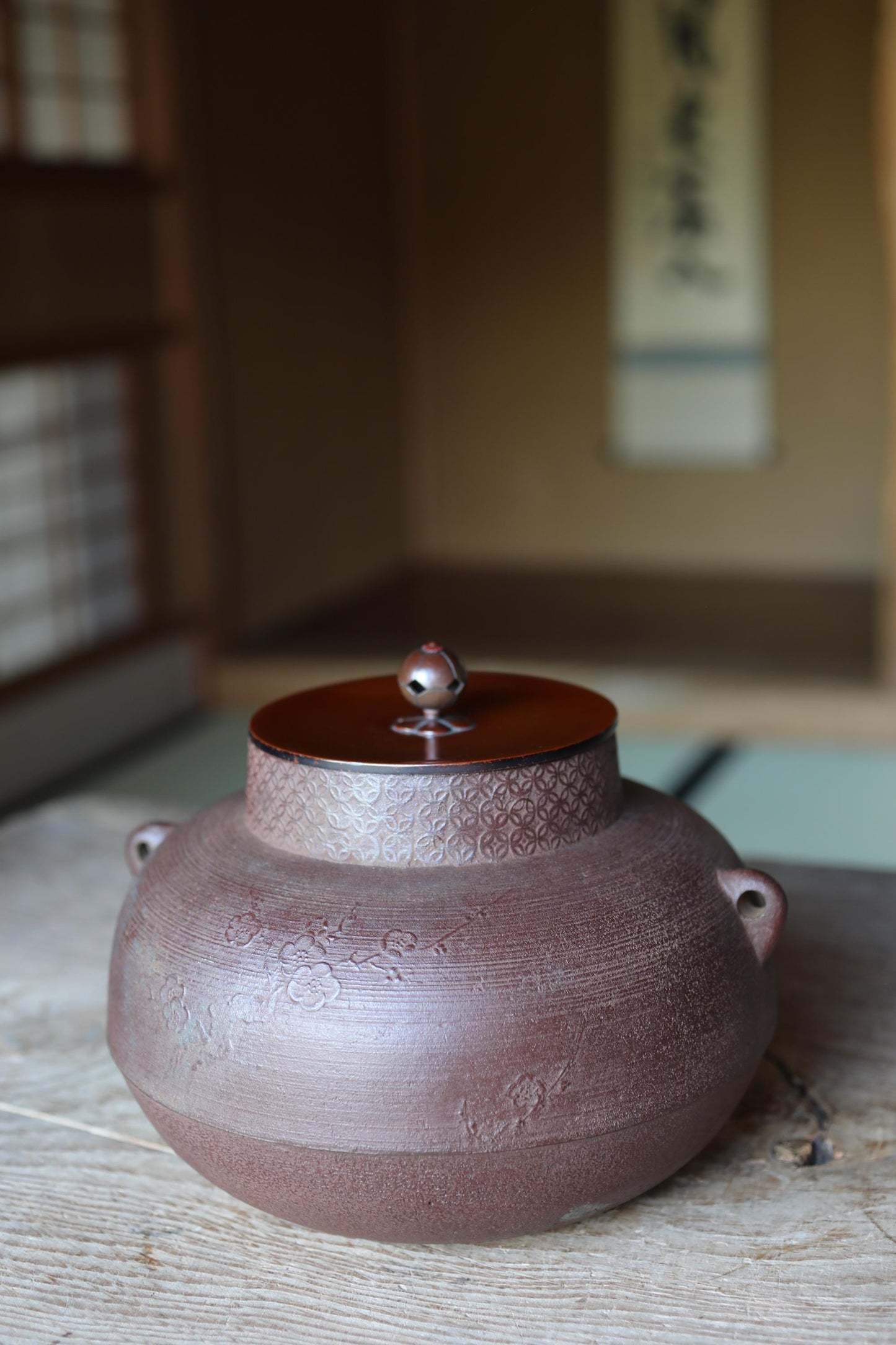 A brown ceramic tea pot with a lid sits on a wooden surface, with a blurred background that includes a window and a wall with Japanese characters.