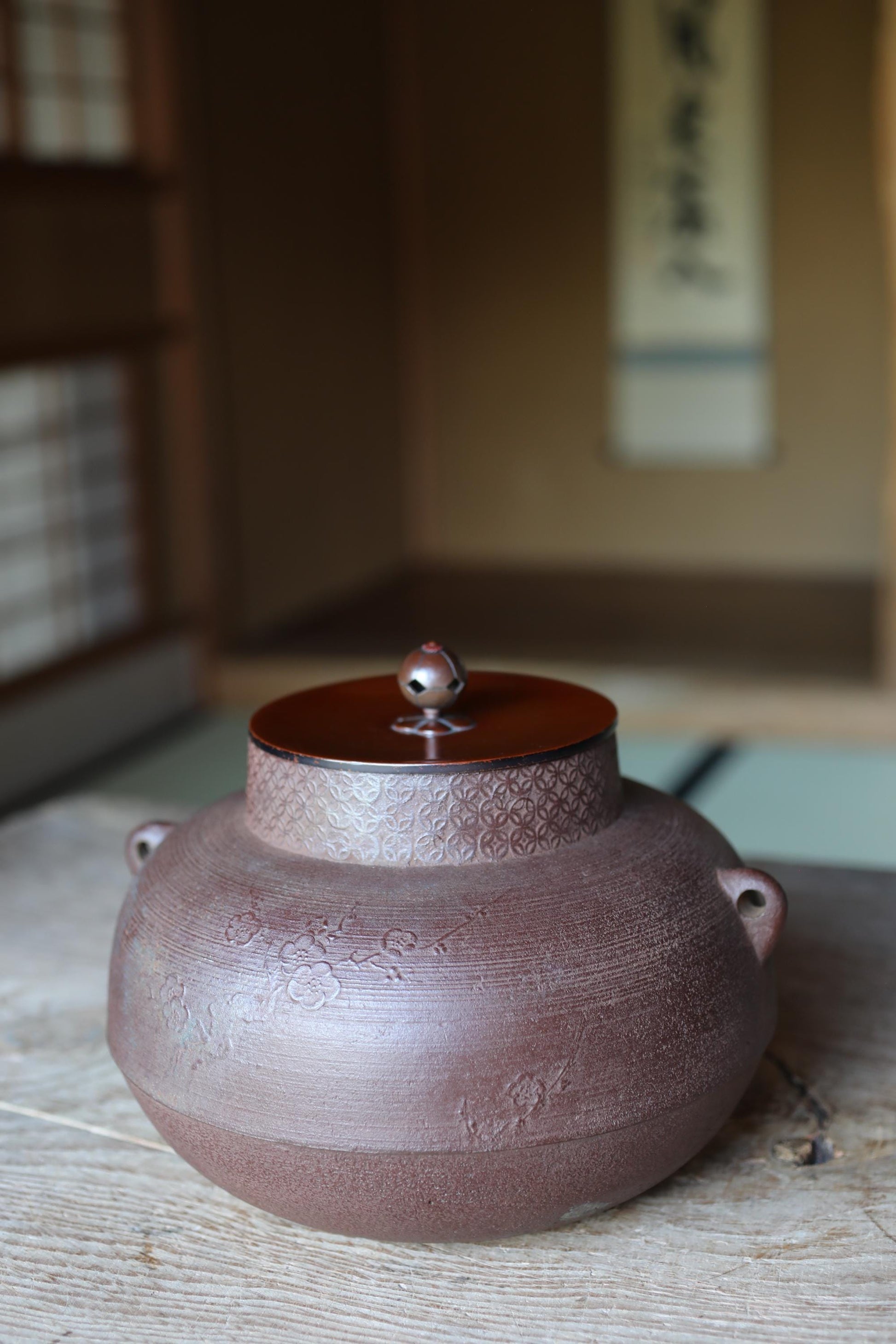 A brown ceramic tea pot with a lid sits on a wooden surface, with a blurred background that includes a window and a wall with Japanese characters.