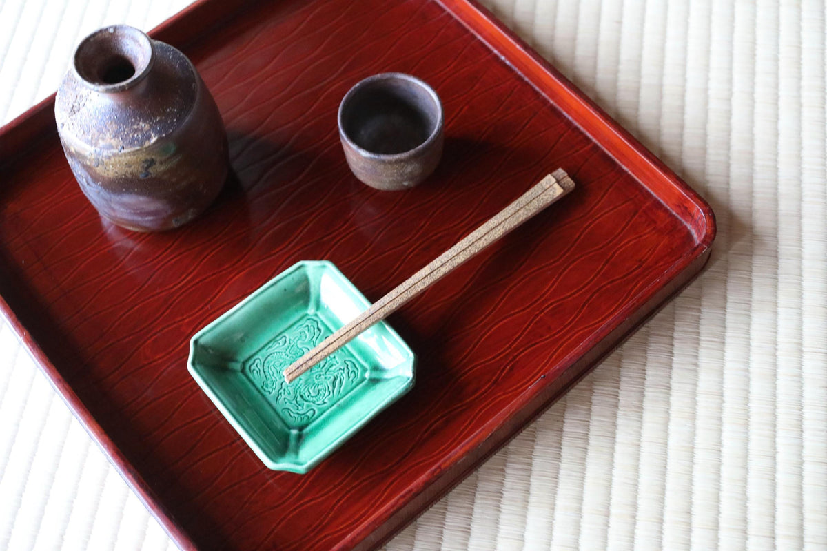A red tray with a green dish, a brown ceramic cup, and a pair of chopsticks on a woven mat.