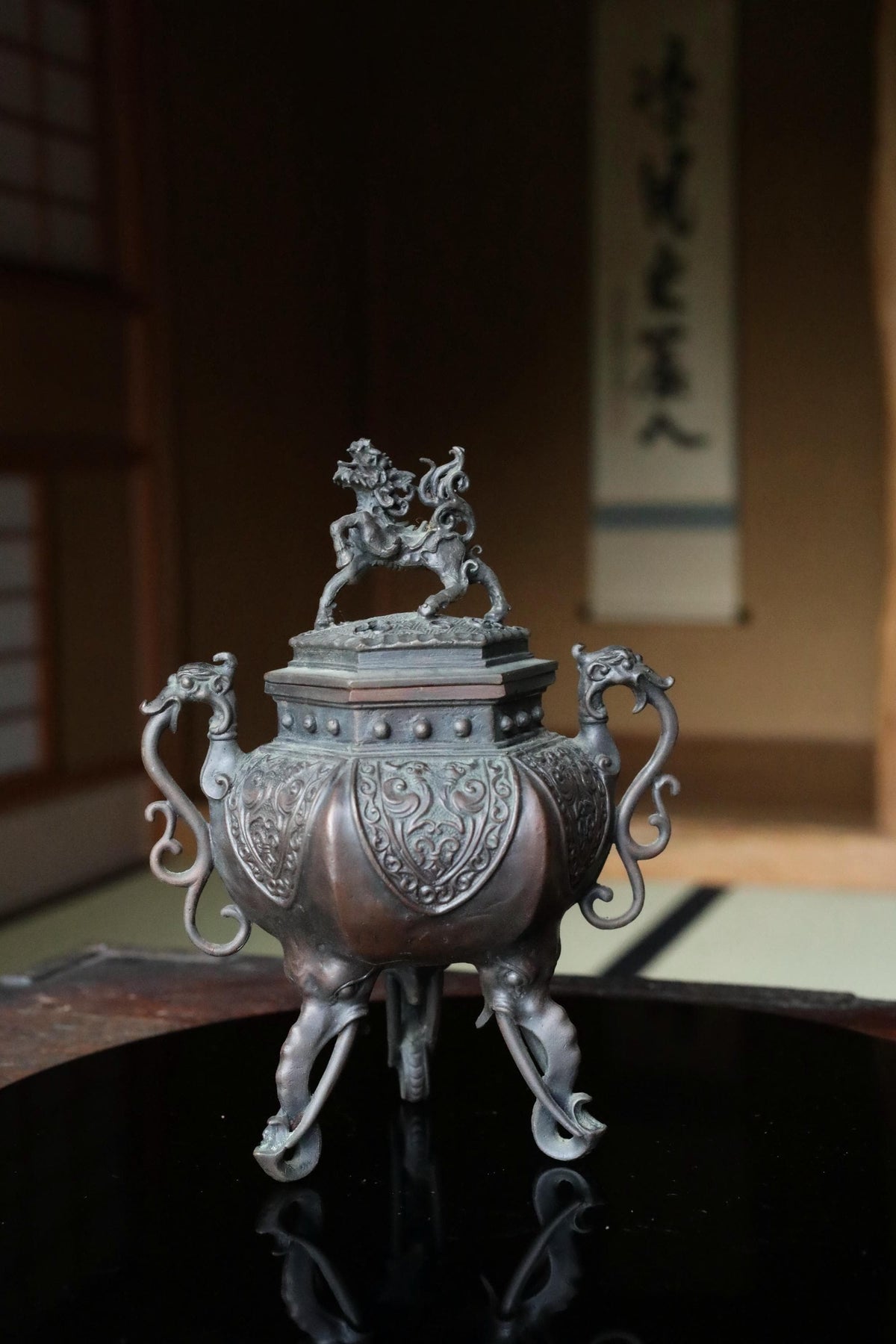 a decorative metal object, possibly a vase or urn, with intricate designs and a dragon figure on top. The object is placed on a reflective surface, possibly a table or countertop.