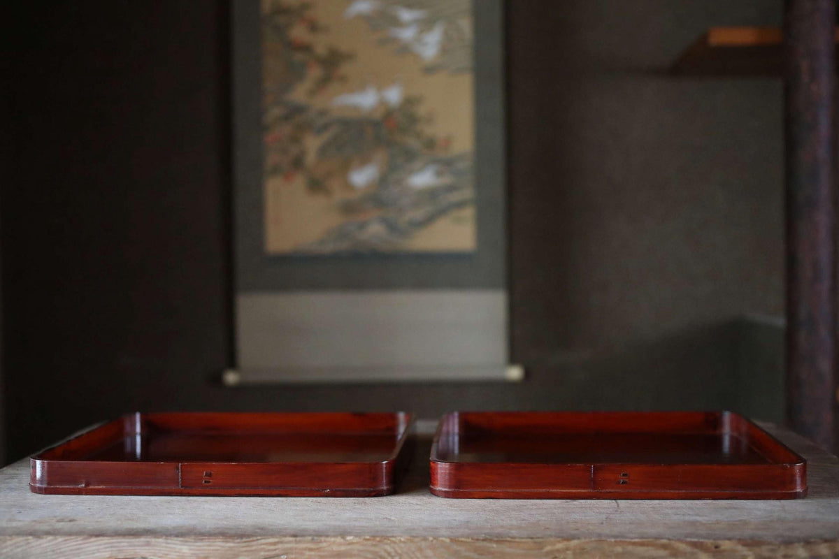 two wooden trays placed on a wooden surface, with a blurred background that includes a painting or artwork.