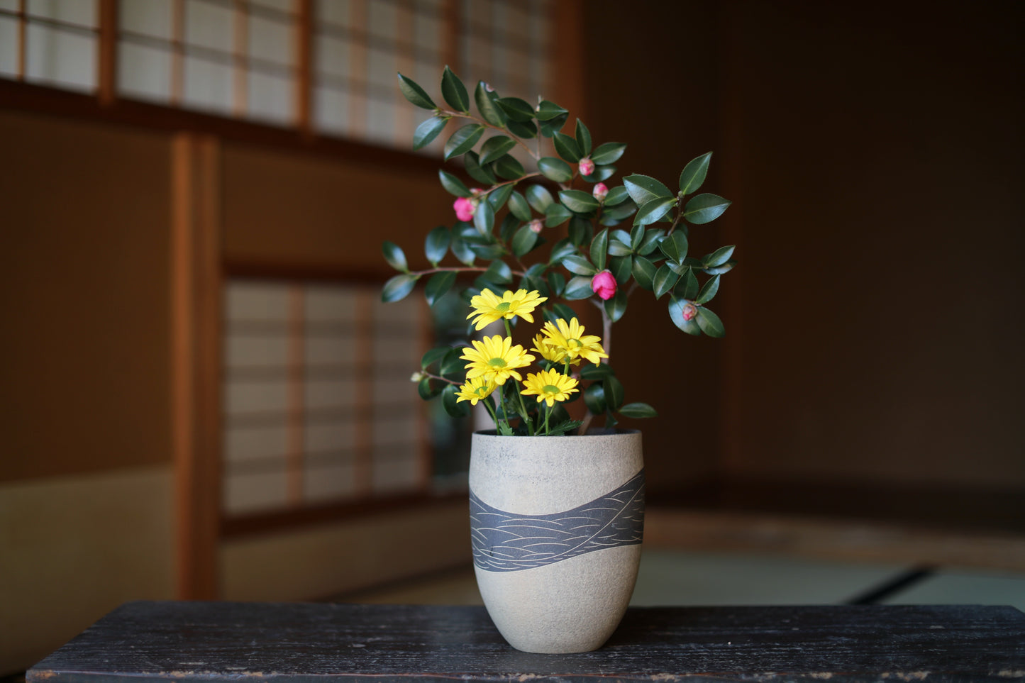 A white vase with a gray stripe holds a bouquet of yellow flowers and green leaves, placed on a wooden surface.
