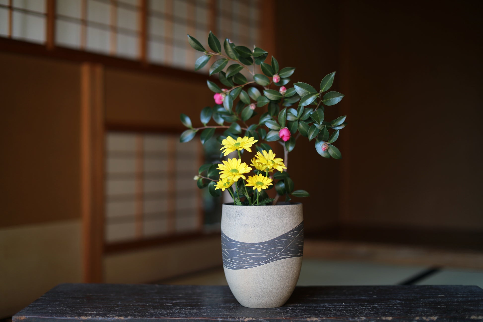 A white vase with a gray stripe holds a bouquet of yellow flowers and green leaves, placed on a wooden surface.