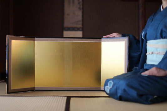 A person in traditional Japanese attire is seated on the floor, holding a golden object that appears to be a decorative item.