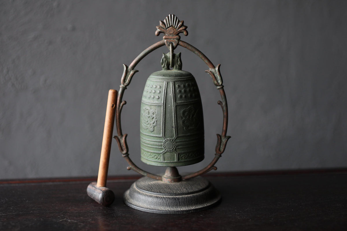 An antique green bell with a decorative metal frame and a wooden mallet is displayed on a dark surface.