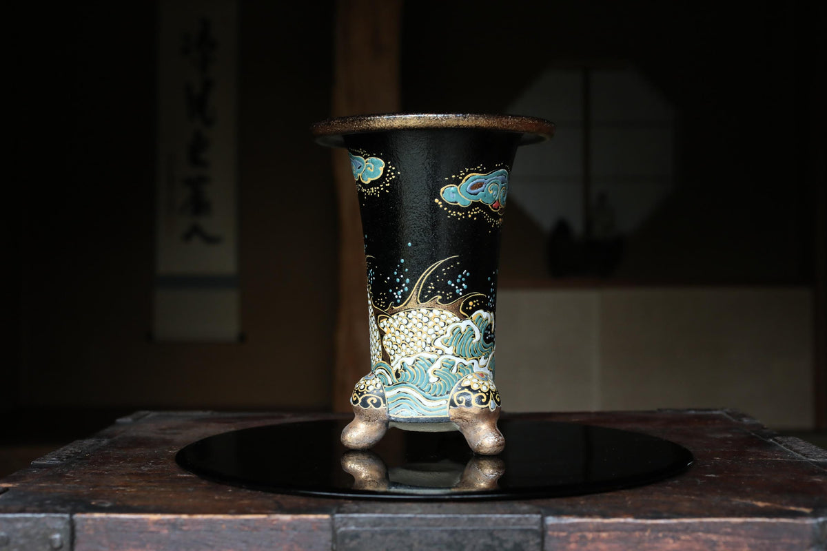 A black vase with intricate blue and gold designs sits on a black circular plate, placed on a wooden surface.