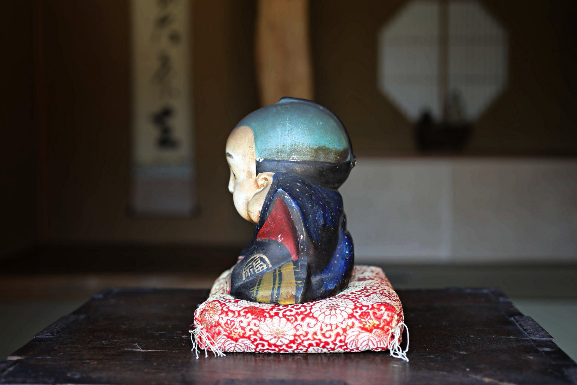 A ceramic figurine of a person wearing a blue hat and a red and blue robe, sitting on a red and white cushion, placed on a wooden surface.