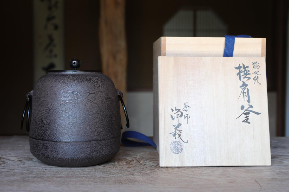 A black ceramic tea kettle with a handle and lid, and a wooden box with Japanese characters on it, both placed on a wooden surface.
