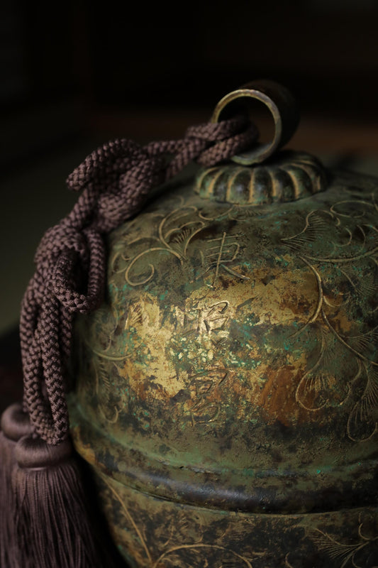 An ornate, antique-looking metal bell or container with a tassel hanging from it, set against a dark background.