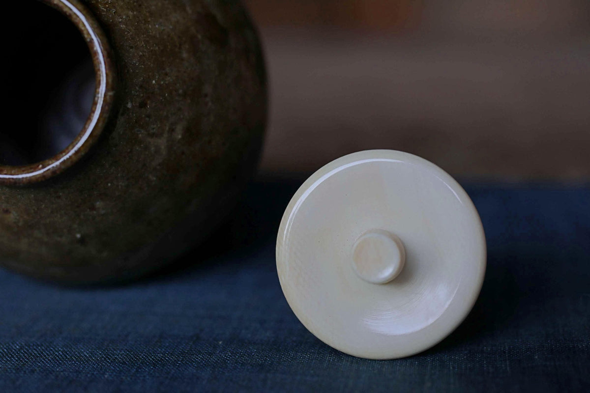 A close-up image of a white ceramic lid on a dark blue surface, with a brown ceramic jar or container visible in the background.
