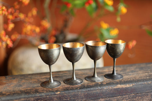 A row of four antique-looking metal wine glasses with gold-colored stems and bowls, placed on a wooden surface against a blurred background of flowers.