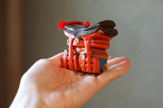 A hand holding a small, red figurine of a temple or shrine with a red tassel on top.