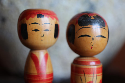 Two wooden dolls with painted faces and red and yellow clothing are shown in the image.