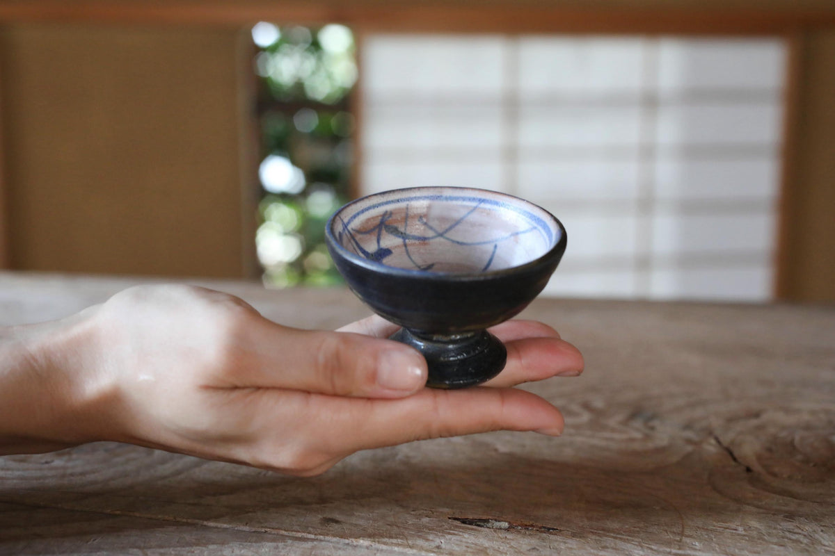 A hand holding a small, black bowl with a blue and white pattern on it, placed on a wooden surface.