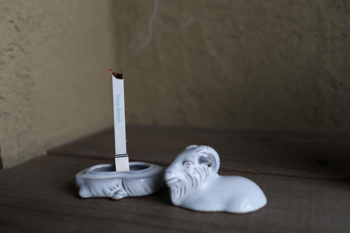 A white ceramic incense burner with a lit incense stick sits on a wooden surface, with smoke rising from the incense.