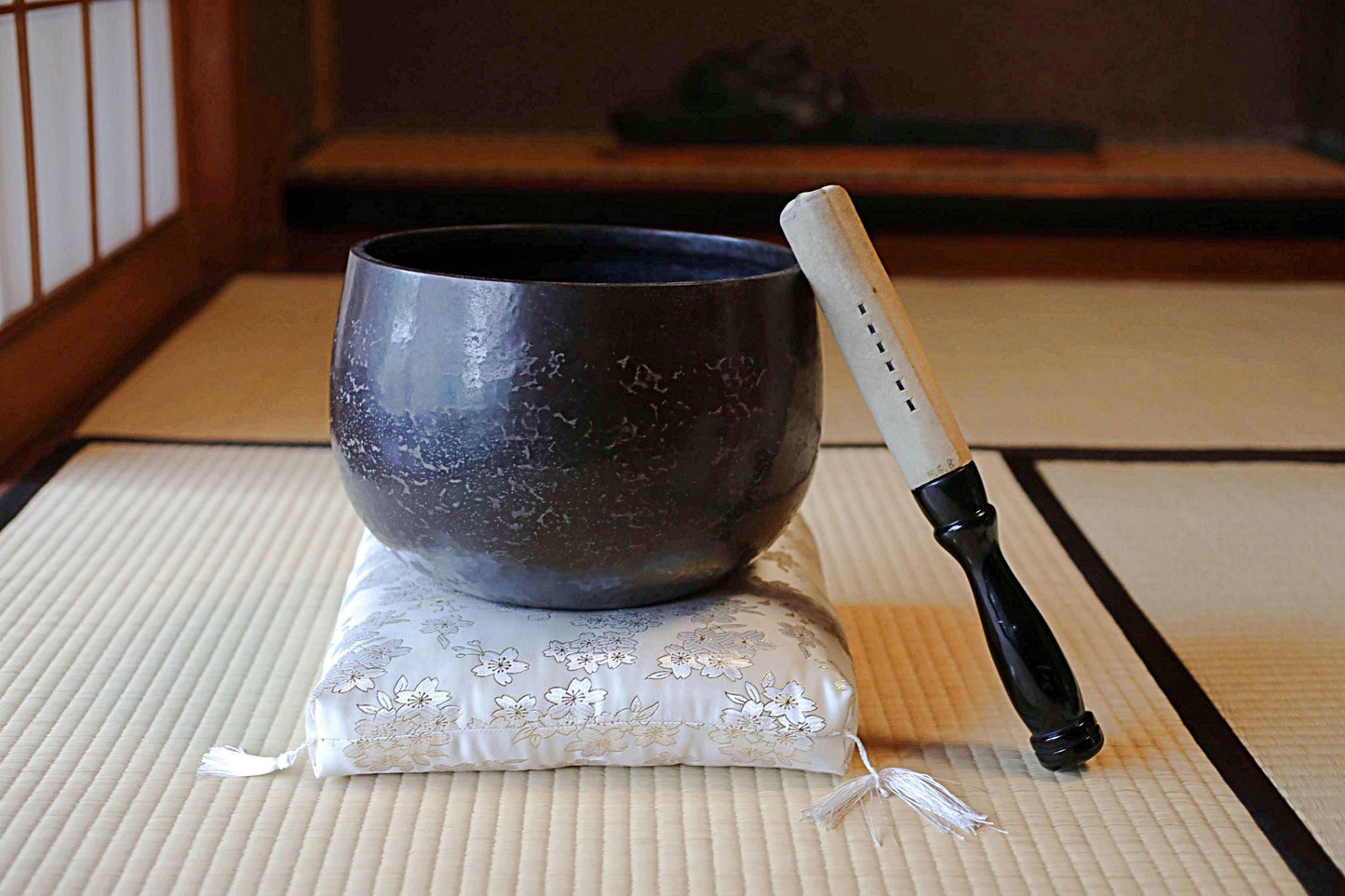 以下が含まれることがあります： 暗い質感のシンギングボウルが、花柄のクッションの上に置かれています。黒い柄の木槌がボウルのそばにあります。畳の上で、瞑想とサウンドヒーリングに焦点を当てたシーンです。