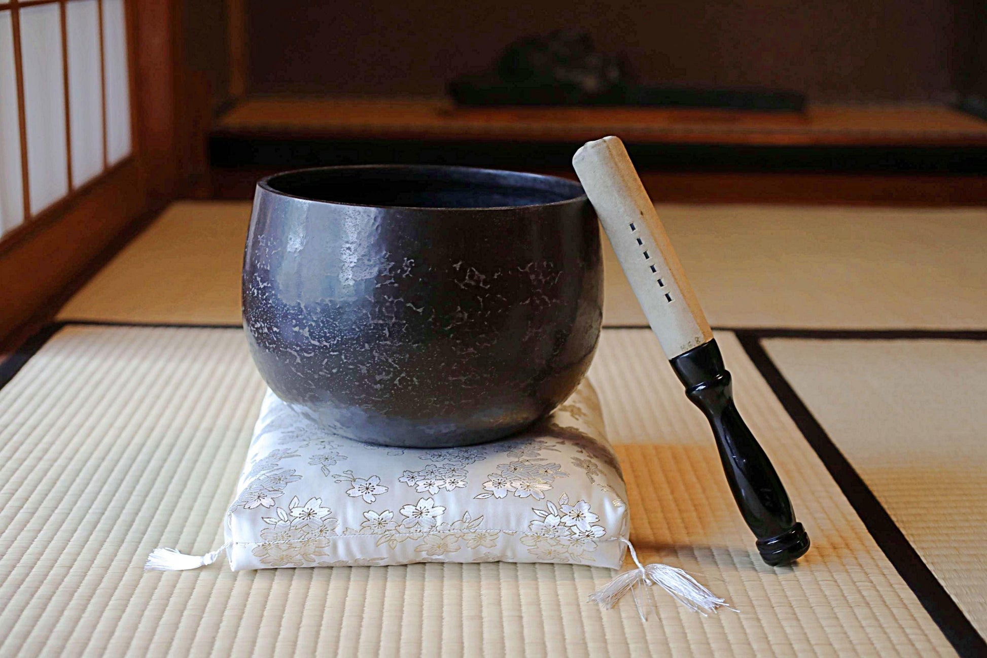 以下が含まれることがあります： 暗い質感のシンギングボウルが、花柄のクッションの上に置かれています。黒い柄の木槌がボウルのそばにあります。畳の上で、瞑想とサウンドヒーリングに焦点を当てたシーンです。