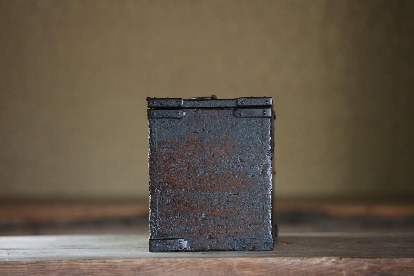 A weathered, black metal box sits on a wooden surface against a plain background.