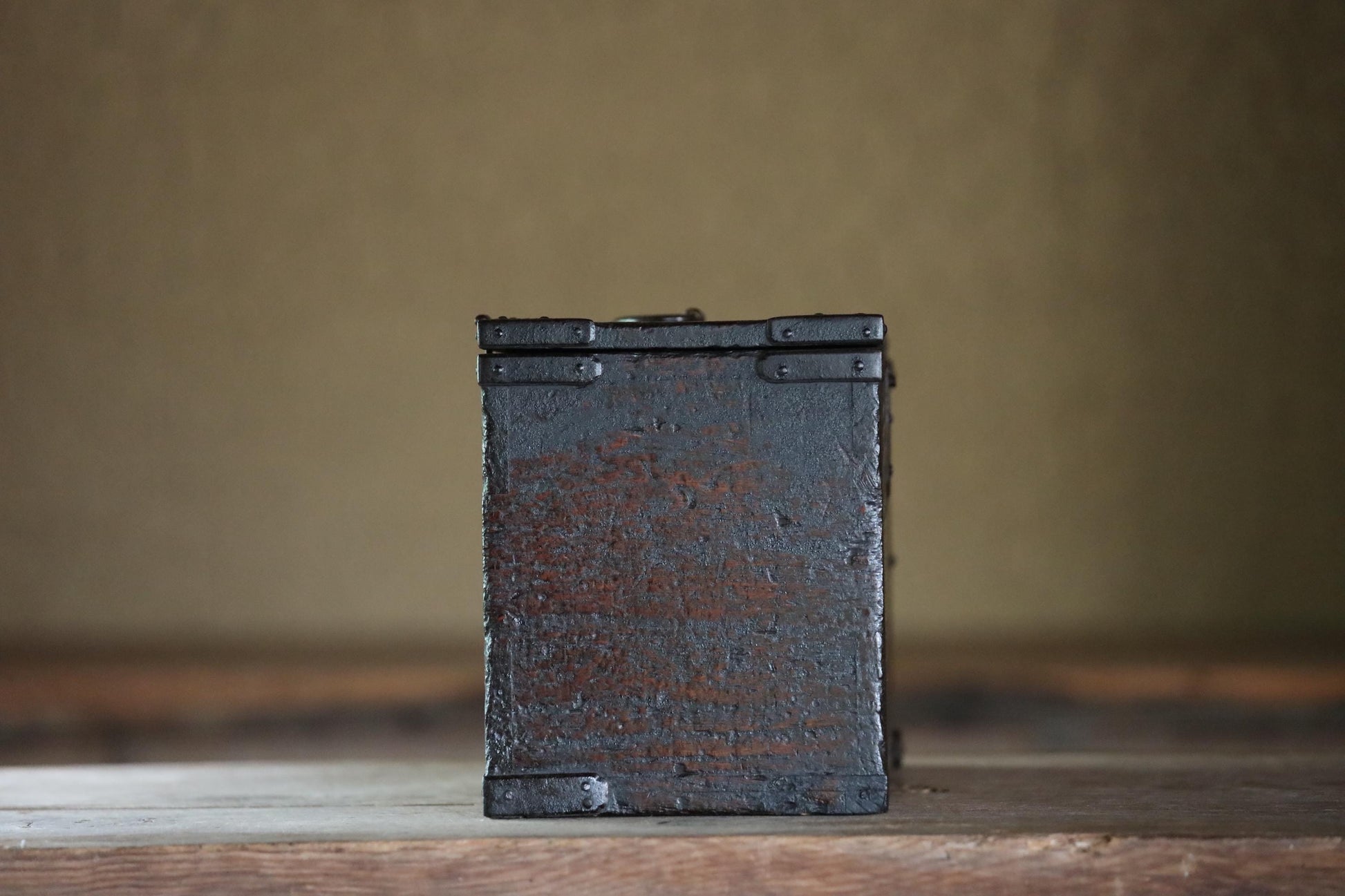 A weathered, black metal box sits on a wooden surface against a plain background.
