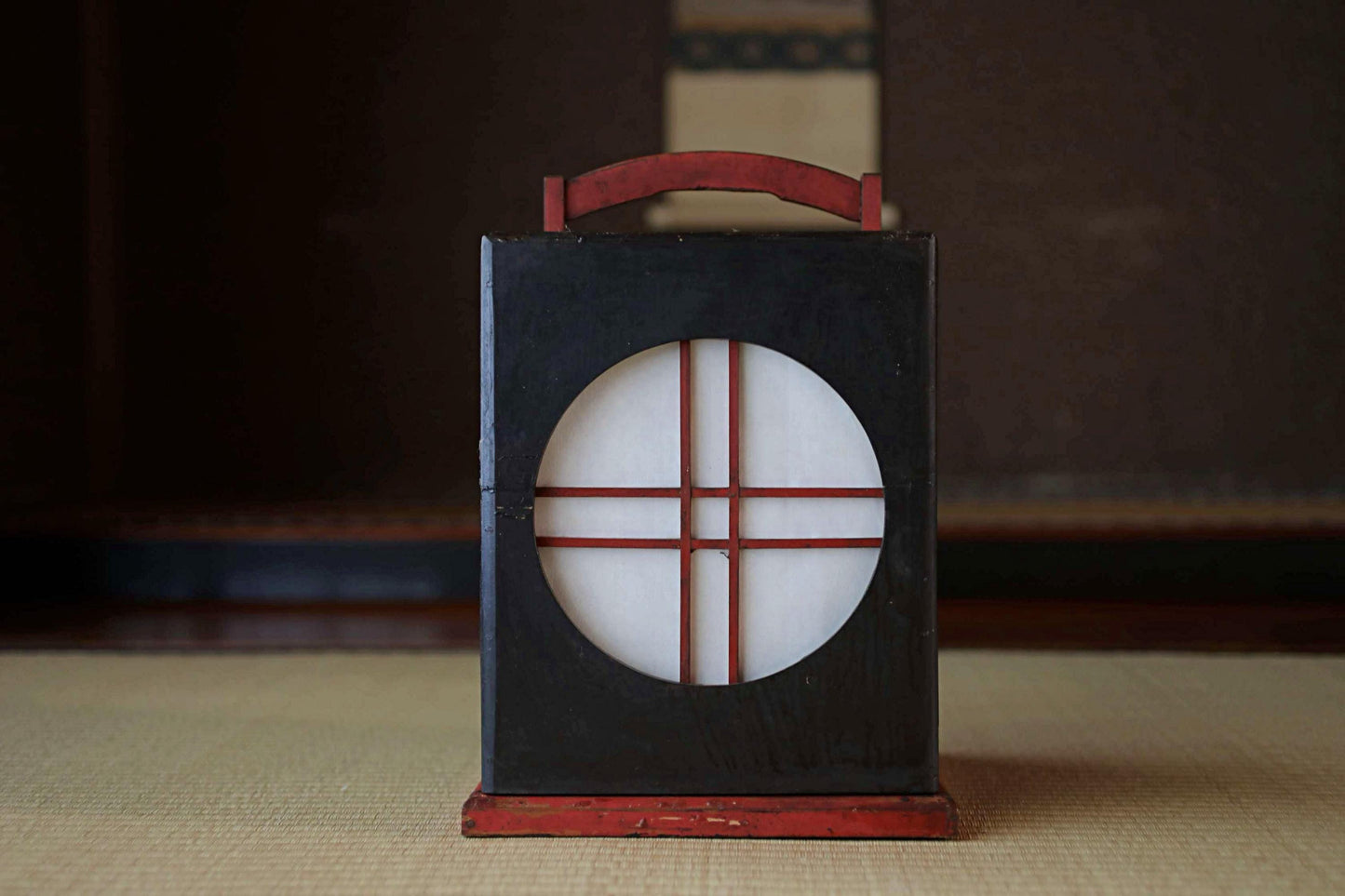 A black lantern with a red handle and a circular design on the front, placed on a beige surface.