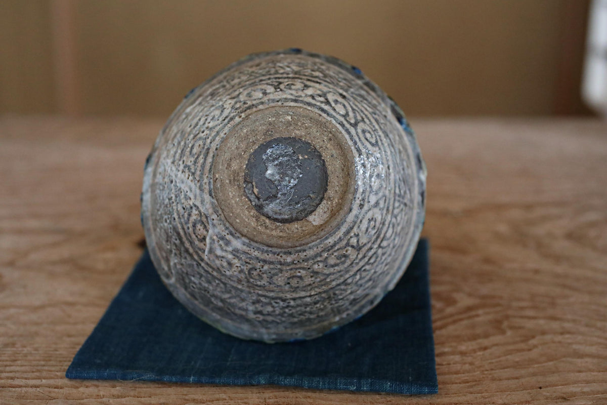 An old, ornate vase with intricate patterns sits on a blue cloth on a wooden surface.