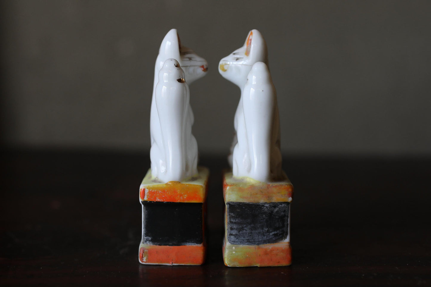 Two white ceramic cat figurines standing on colorful bases against a dark background.