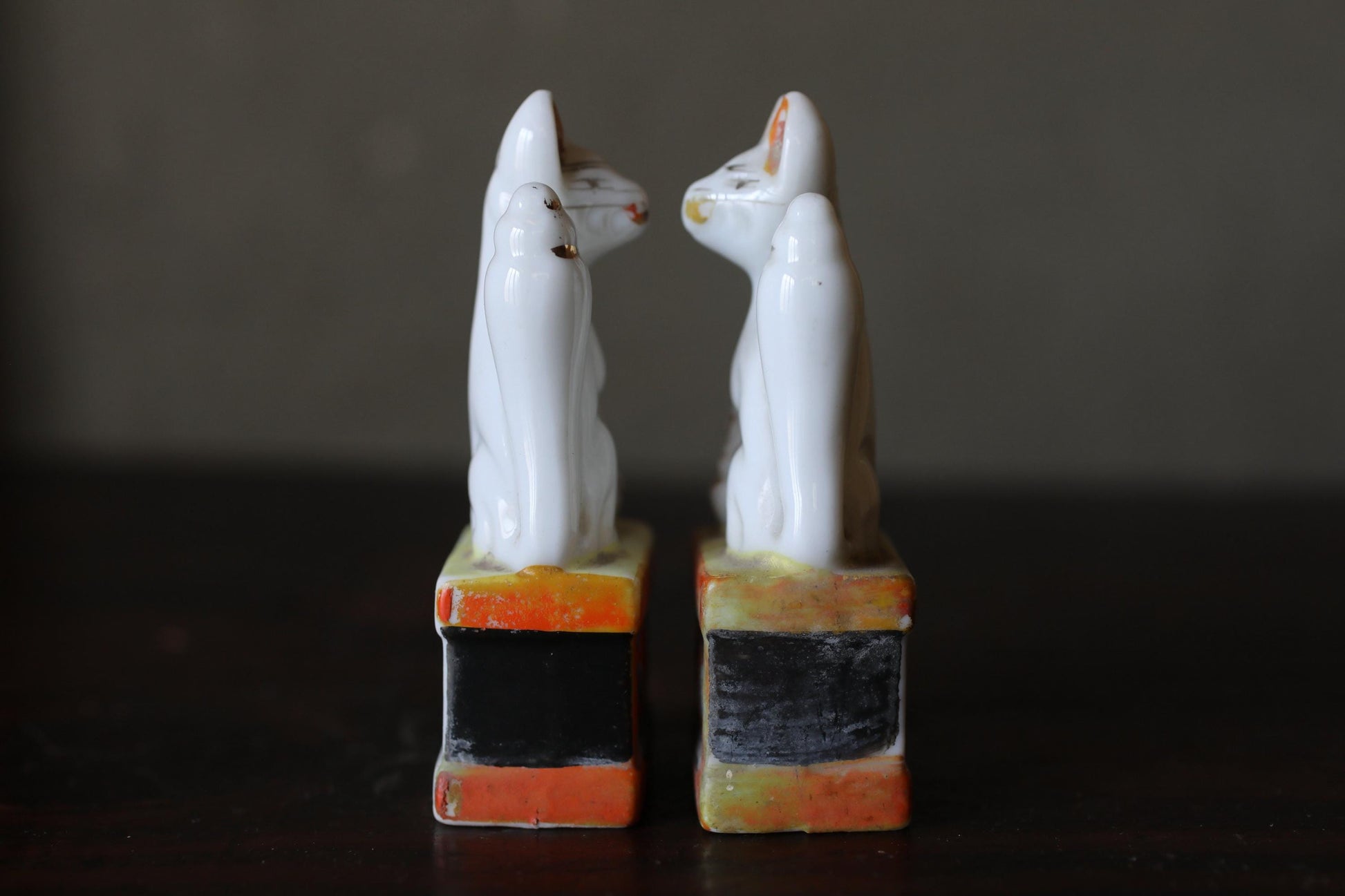 Two white ceramic cat figurines standing on colorful bases against a dark background.