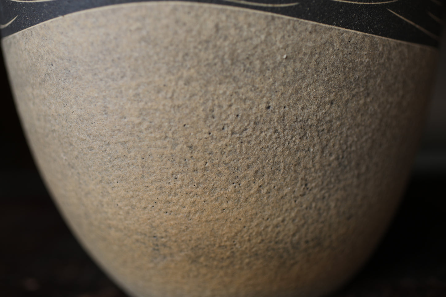A close-up view of a textured, beige-colored ceramic object, possibly a vase or bowl, with a dark border around the edge.
