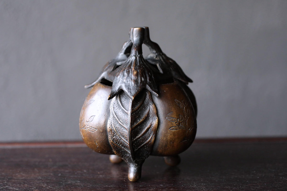 A bronze incense burner with a leaf-like design on the front sits on a dark wooden surface.