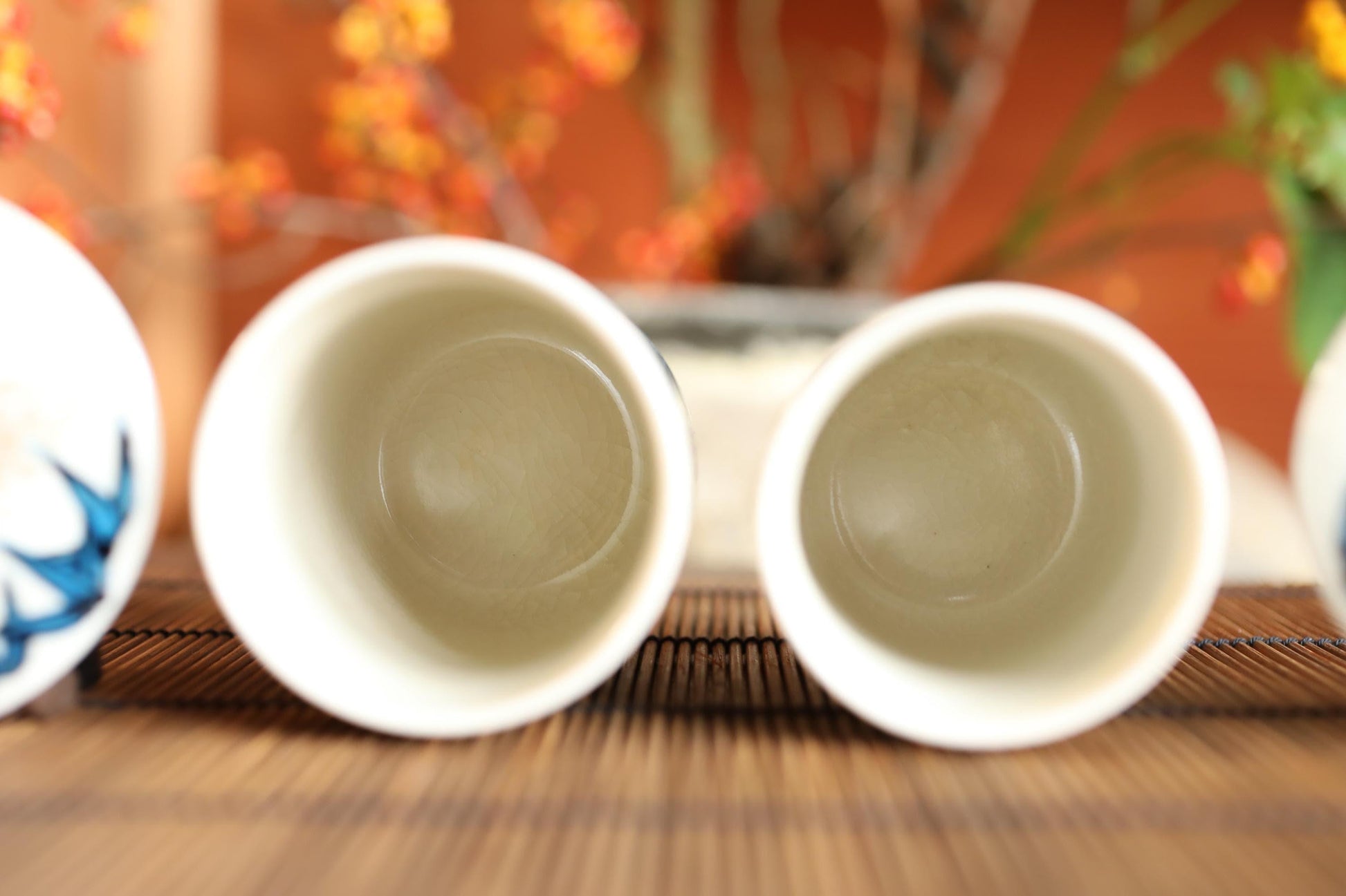 Two white cups with blue designs on them are placed on a bamboo mat, with a blurred background featuring orange and yellow flowers.
