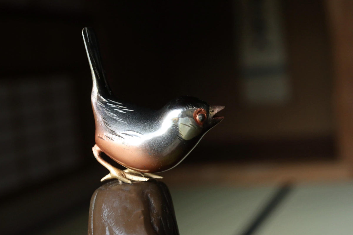A small, shiny bird figurine perched on a brown object.